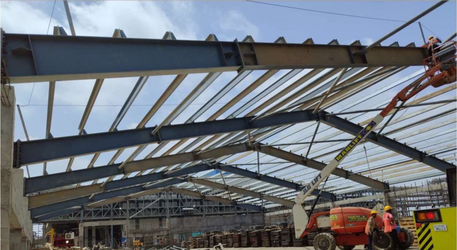 A pre-engineered steel building frame being erected on site, with rafters set and a manlift alongside