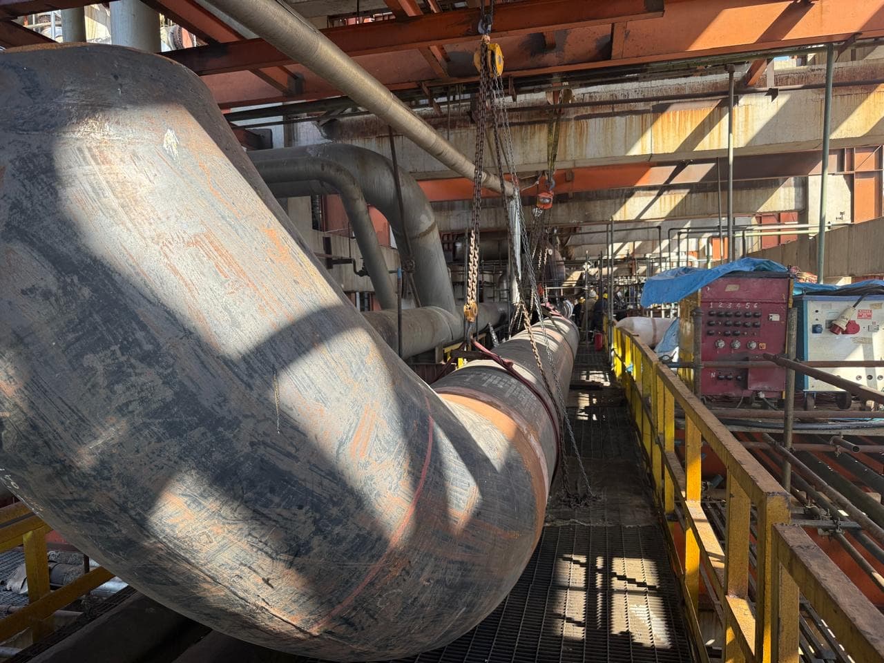 Large-bore process piping and elbows being rigged inside a plant, on elevated grating