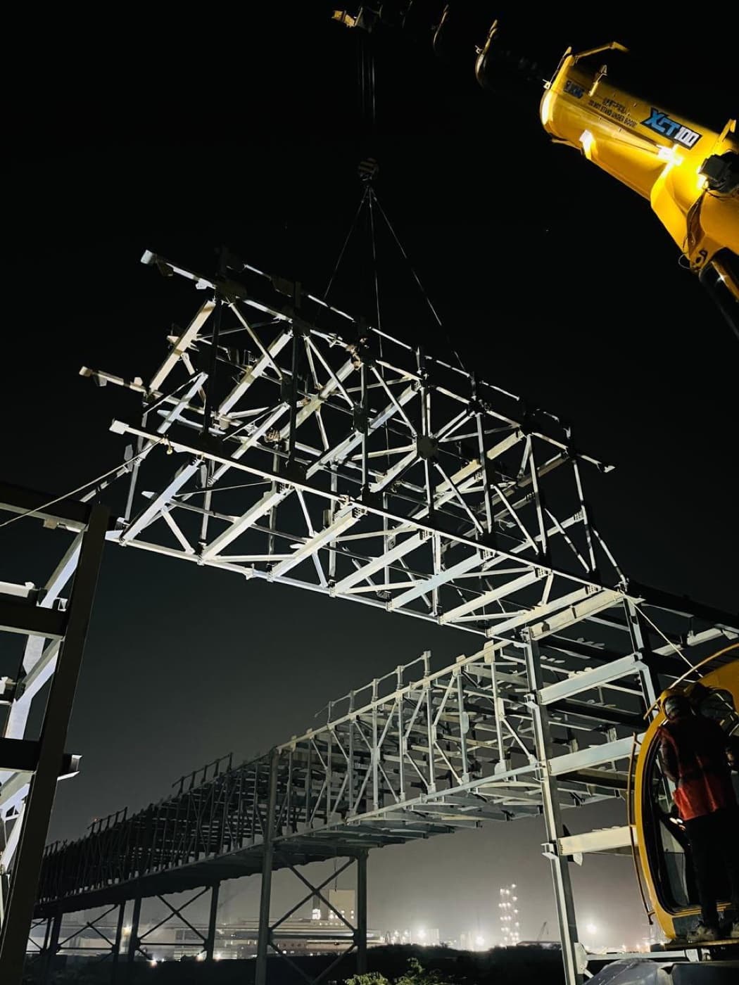 A fabricated steel roof truss being lifted into place by crane at night